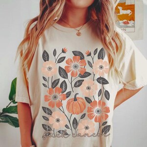 May include: Cream-coloured t-shirt with a floral design. Peach and light pink flowers, grey leaves, and a pumpkin are featured. The words "fuck cancer" are written in a script font at the bottom of the shirt.