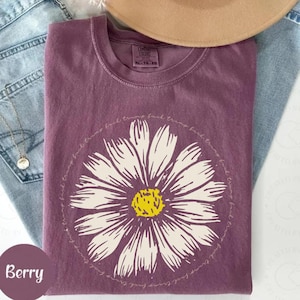 May include: A purple t-shirt with a white daisy graphic and the text "back, trust, back, trust, back, trust, back, trust" repeated around the daisy. The shirt is folded and laid on a pair of blue jeans.