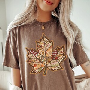 May include: Brown t-shirt with a large maple leaf design. The leaf is filled with a patchwork of autumn patterns and the text "Fuck Trump". The shirt has short sleeves and a relaxed fit.