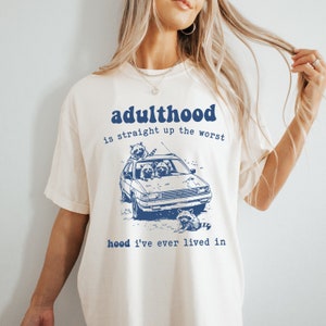May include: A white t-shirt with a blue graphic print of raccoons in a car. The text on the shirt reads "adulthood is straight up the worst hood I've ever lived in".