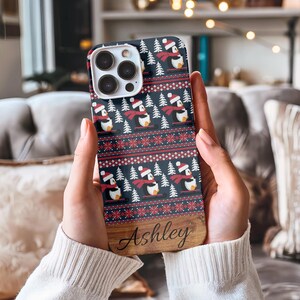 May include: A personalized phone case with a wintery design featuring penguins wearing Santa hats and scarves. The case has a wood-grain background with the name "Ashley" printed on it.