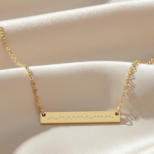 May include: A gold-colored necklace with a rectangular pendant. The pendant features a series of dots and dashes, likely representing a message in Morse code. The chain and pendant are delicate, set against a soft, off-white fabric background.