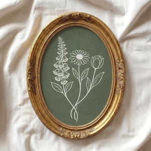 May include: An oval, gold-colored framed print featuring a botanical illustration. The artwork showcases white line drawings of a foxglove, daisy, and tulip on a muted green background. The frame has ornate detailing.