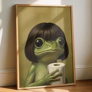 May include: A framed art print featuring a whimsical frog with large eyes and a dark bob haircut. The frog is holding a roll of toilet paper. The artwork has a vintage aesthetic with a muted color palette and is displayed in a wooden frame.