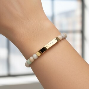 May include: A beaded bracelet with a gold bar. The bracelet features a series of small, round, light-colored beads. The gold bar in the center has a series of dots and dashes, possibly Morse code.