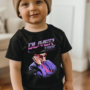 May include: A black short-sleeved t-shirt featuring a graphic print of a person wearing red sunglasses and a purple jacket. The text OLIVER TREE is printed in a stylized purple and white font above the person. The shirt has a crew neckline.