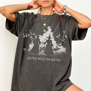 May include: Dark grey t-shirt with a graphic print of witches around a bonfire, with the text "girls will be girls" in a white font. The shirt has short sleeves and a relaxed fit.