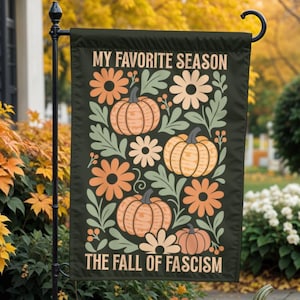 May include: A black garden flag with the text "MY FAVORITE SEASON THE FALL OF FASCISM" and a design of pumpkins and flowers in orange, cream, and green. Three pumpkins are in front of the flag, with fall foliage in the background.