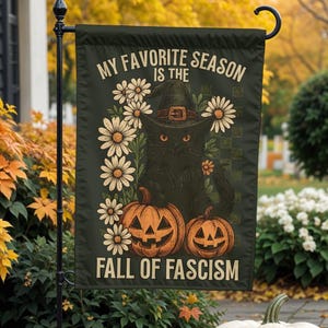 May include: A dark green garden flag with the text "MY FAVORITE SEASON IS THE FALL OF FASCISM". The flag features a black cat wearing a witch hat, surrounded by daisies and jack-o'-lanterns. Pumpkins and fall foliage are in the foreground.