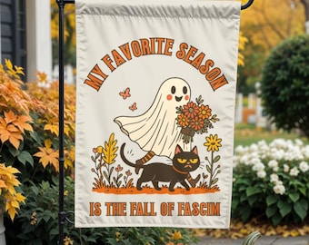 May include: A beige garden flag with the text "MY FAVORITE SEASON IS THE FALL OF FASCISM" in orange. The flag features a cartoon ghost, black cat, flowers, and butterflies in a fall theme. Pumpkins and foliage are in the background.