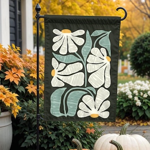 May include: A black garden flag featuring a floral design with white flowers, light green leaves, and yellow centers. The words "FUCK TRUMP" are printed on the leaves. The flag is displayed outdoors with pumpkins and fall foliage in the background.