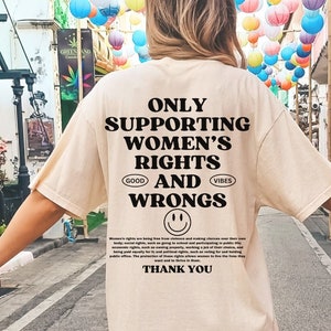 May include: A beige t-shirt with black text that reads "Only Supporting Women's Rights and Wrongs." The text is in a bold, sans-serif font. There is a smiley face below the text. The shirt is being worn by a person walking down a street.