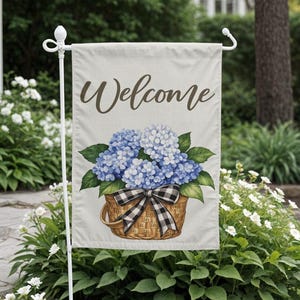 May include: A garden flag with the word "Welcome" in brown script. The flag features a watercolour illustration of blue hydrangeas in a woven basket with a black and white chequered bow. The flag is displayed on a white metal stand.
