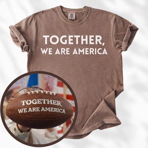 Könnte beinhalten: Braunes T-Shirt mit dem weißen Text "TOGETHER, WE ARE AMERICA". Ein Football mit demselben Text befindet sich in einem kreisförmigen Einsatz. Das Shirt hat hochgekrempelte Ärmel und einen Rundhalsausschnitt.