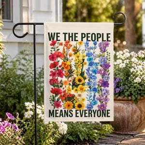 May include: A garden flag with the text "WE THE PEOPLE MEANS EVERYONE" in black. The flag features a vibrant floral design in a rainbow of colors, including red, orange, yellow, green, blue, and purple flowers.