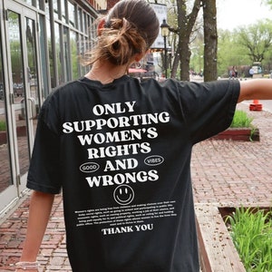 May include: Black t-shirt with white text that reads "Only Supporting Women's Rights and Wrongs." The text is surrounded by a smiley face and the words "Good Vibes".