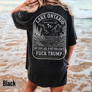 May include: A black short-sleeve t-shirt featuring a white graphic on the back. The design depicts a lighthouse, trees, and a sailboat on Lake Ontario with the text: LAKE ONTARIO, PROTECT THE GREAT LAKES, NOT YOUR LAKE. NOT YOUR LEGACY. FUCK TRUMP.