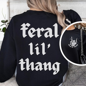 Może przedstawiać: Czarna bluza z napisem "feral lil' thang" w białym, gotyckim kroju pisma na plecach. Mniejszy obrazek po prawej stronie przedstawia gest dłoni z tym samym tekstem.