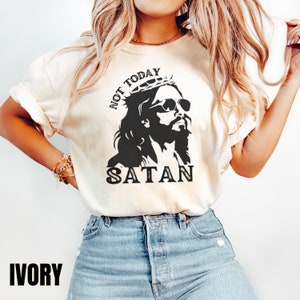 Pu&ograve; includere: Una maglietta bianca con una grafica nera di Ges&ugrave; che indossa occhiali da sole e una corona. Il testo "NOT TODAY SATAN" &egrave; stampato sulla maglietta.