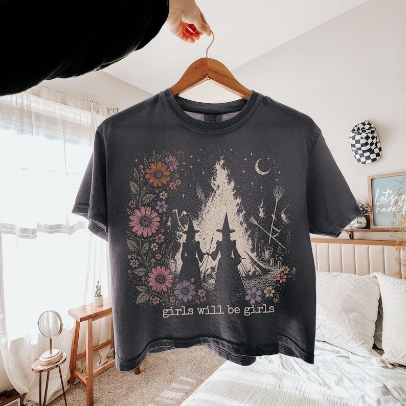 May include: Dark gray t-shirt featuring a graphic of two witches holding hands in front of a bonfire, surrounded by flowers and a crescent moon. The text "girls will be girls" is printed at the bottom of the shirt.