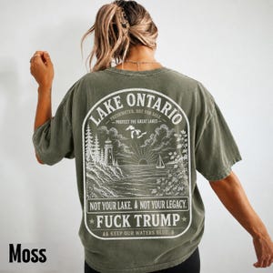 May include: An olive green t-shirt featuring a white graphic on the back. The design depicts a lighthouse, a sailboat, and a sun over Lake Ontario. Text includes "LAKE ONTARIO," "FRESHWATER. NOT FOR SALE," "PROTECT THE GREAT LAKES," and "FUCK TRUMP."