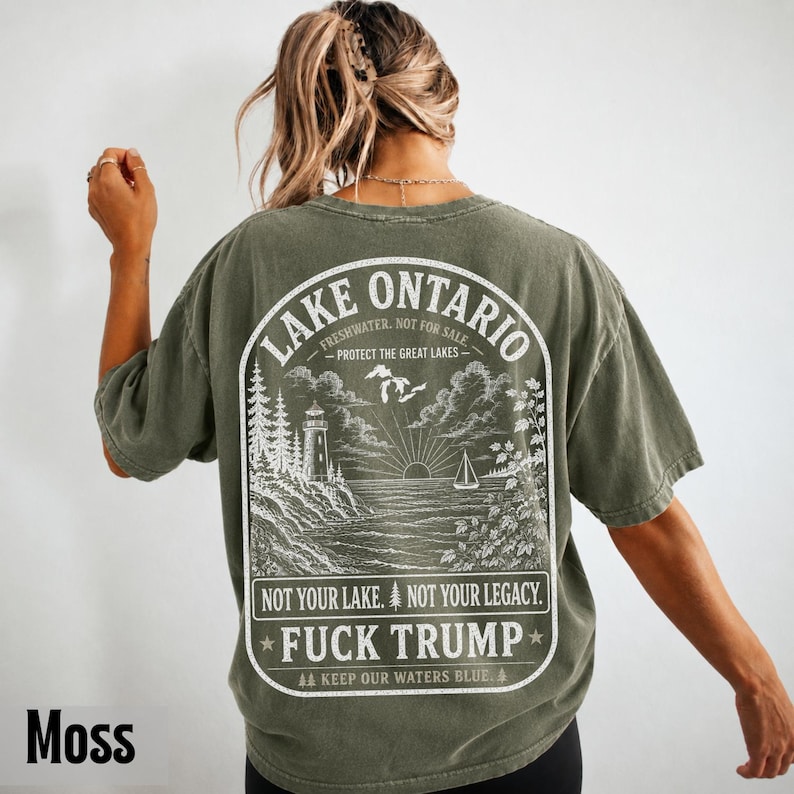 May include: An olive green t-shirt featuring a white graphic on the back. The design depicts a lighthouse, a sailboat, and a sun over Lake Ontario. Text includes "LAKE ONTARIO," "FRESHWATER. NOT FOR SALE," "PROTECT THE GREAT LAKES," and "FUCK TRUMP."