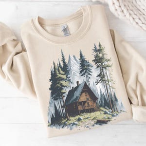 May include: A beige sweatshirt featuring a mountain cabin scene. The graphic depicts a wooden cabin nestled among tall pine trees and snow-capped mountains. The design is rendered in shades of green, gray, and brown, creating a nature-inspired aesthetic.