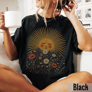 Puede incluir: Camiseta negra con un gr&aacute;fico de sol con los ojos cerrados y un dise&ntilde;o de pradera floral. El sol tiene rayos dorados y una cara melocot&oacute;n, rodeada de flores coloridas. La palabra "Black" est&aacute; impresa en la parte inferior derecha.