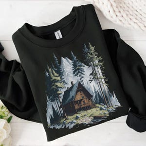 May include: Black sweatshirt featuring a mountain cabin scene. The graphic depicts a wooden cabin nestled among tall pine trees and snow-capped mountains. The design is in shades of green, gray, and brown.