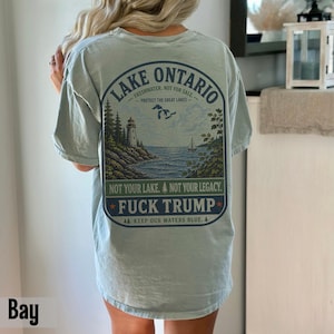 May include: A light blue t-shirt featuring a graphic on the back with a lighthouse, trees, and a sailboat on Lake Ontario. The text reads: LAKE ONTARIO, FRESHWATER, NOT FOR SALE, PROTECT THE GREAT LAKES, NOT YOUR LAKE, NOT YOUR LEGACY, FUCK TRUMP.