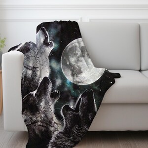 May include: A black and white blanket featuring a nighttime scene with three wolves howling at a full moon. The blanket has a dark background with stars and a nebula effect, creating a dramatic and nature-inspired design.
