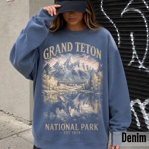 May include: Denim blue sweatshirt featuring a scenic illustration of the Grand Teton National Park. The design includes mountains, a lake, and trees, with the text "GRAND TETON NATIONAL PARK - EST 1929". The sweatshirt is a casual style.