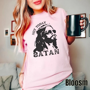 Pu&ograve; includere: Maglietta rosa con una grafica in bianco e nero di Ges&ugrave; con occhiali da sole e una corona con il testo "Not Today Satan".