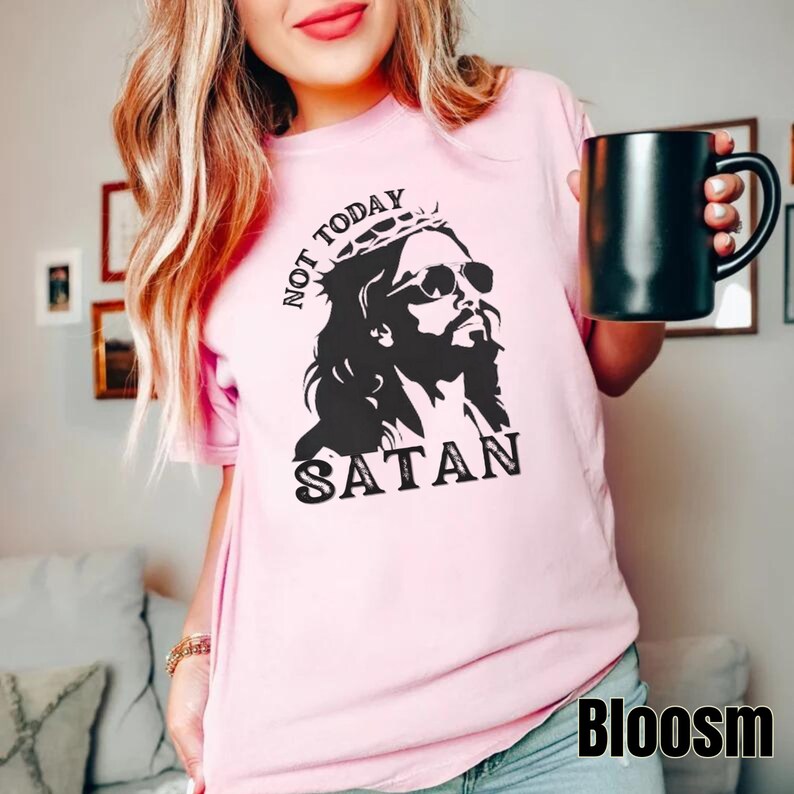 Pu&ograve; includere: Maglietta rosa con una grafica in bianco e nero di Ges&ugrave; con occhiali da sole e una corona con il testo "Not Today Satan".