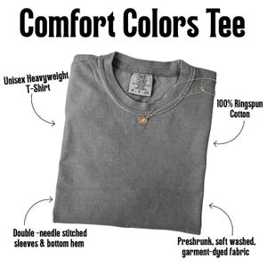 May include: A charcoal gray t-shirt with a crew neckline and a Comfort Colors brand label at the collar. The shirt features double-needle stitched sleeves and bottom hem, made of preshrunk, soft washed, garment-dyed, 100% ringspun cotton fabric.