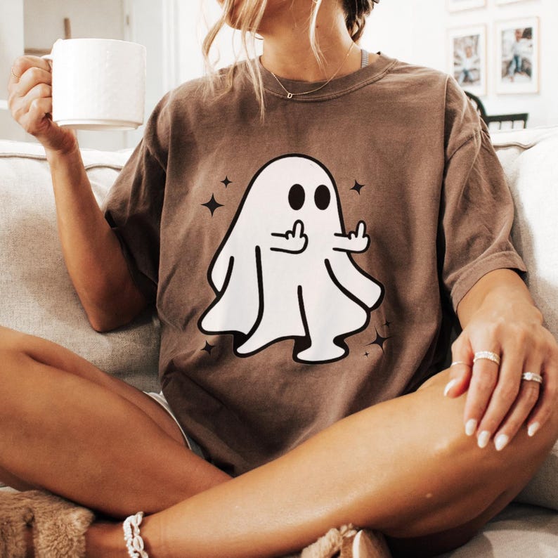 May include: Brown t-shirt featuring a white ghost graphic with two black eyes and a hand gesture. The ghost is surrounded by star shapes. The shirt is worn by a person sitting on a couch.
