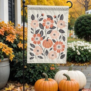 Puede incluir: Una bandera de jard&iacute;n color crema con un dise&ntilde;o floral y de calabaza en tonos melocot&oacute;n, coral y gris. La bandera se exhibe al aire libre con tres calabazas al frente, creando un tema oto&ntilde;al.