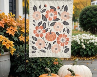 May include: A cream-colored garden flag featuring a floral and pumpkin design in shades of peach, coral, and gray. The flag is displayed outdoors with three pumpkins in front, creating a fall theme.