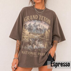May include: Espresso-colored oversized t-shirt featuring a scenic illustration of the Grand Teton National Park. The graphic includes mountains, a lake, and trees, with the text "GRAND TETON NATIONAL PARK - EST 1929-". The shirt has short sleeves.