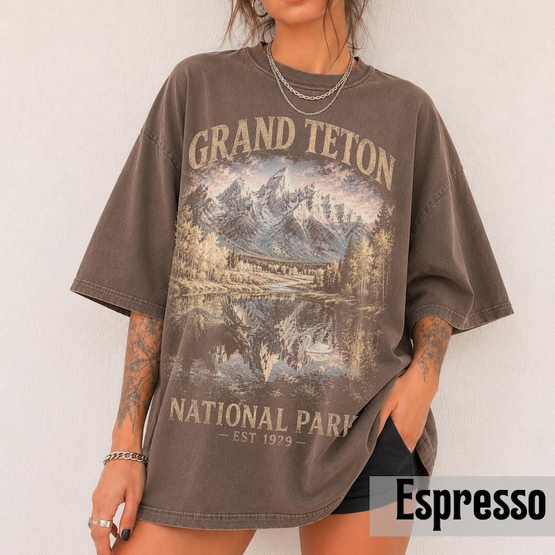 May include: Espresso-colored oversized t-shirt featuring a scenic illustration of the Grand Teton National Park. The graphic includes mountains, a lake, and trees, with the text "GRAND TETON NATIONAL PARK - EST 1929-". The shirt has short sleeves.
