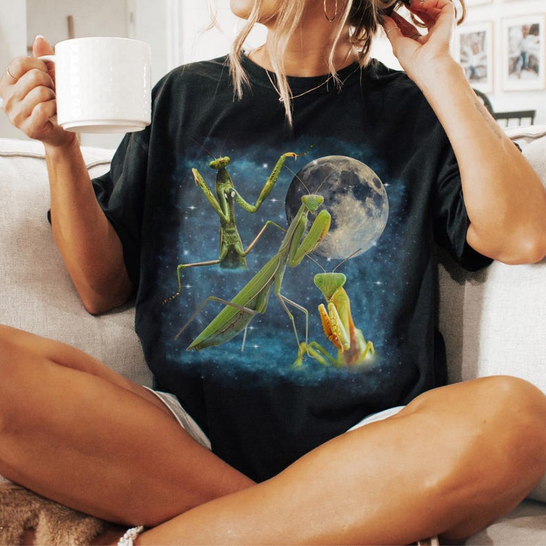 Puede incluir: Camiseta negra con un gr&aacute;fico detallado de mantis religiosas, una luna llena y un cielo estrellado. El dise&ntilde;o presenta mantis religiosas verdes y amarillas sobre un fondo azul oscuro, creando un efecto visual llamativo.