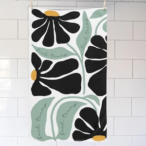 May include: White towel with a floral design featuring large black flowers with yellow centers and light green leaves. The words "fuck trump" are written in a script font on the leaves. The towel is hanging on a clothesline.