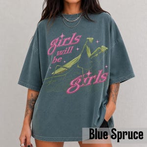 以下が含まれることがあります： チャコールグレーのオーバーサイズTシャツで、緑色のカマキリの大きなグラフィックが特徴です。胸元にはピンクの縁取り文字で「girls will be girls」とプリントされており、デザイン全体に小さなピンクの星が散りばめられています。