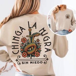 May include: Beige sweatshirt with a graphic on the back featuring a snake coiled around a sword, surrounded by flowers. The text reads "CHINGA LA MIGRA SIN MIEDO". A smaller graphic is on the front, with the same text.