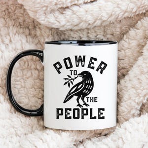 May include: White ceramic mug with a black handle and rim. The mug features the text "POWER TO THE PEOPLE" in a distressed font, along with a black illustration of a bird holding a branch. The mug is set against a soft, textured background.