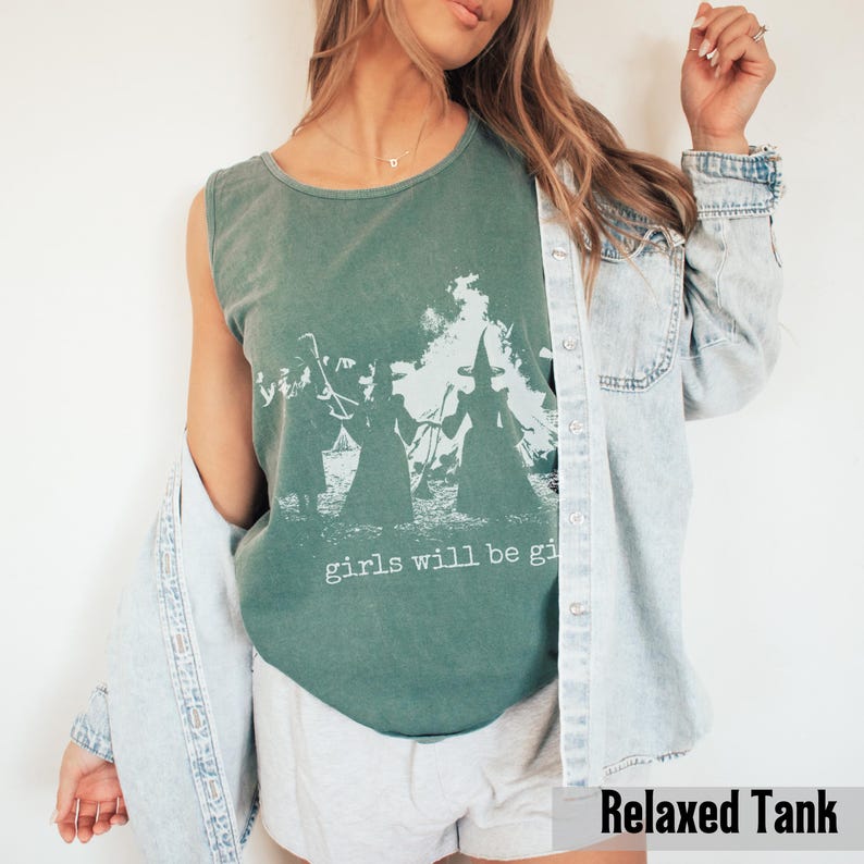 Pu&ograve; includere: Una canotta verde salvia senza maniche con una stampa grafica bianca di due streghe davanti a un fal&ograve; e la scritta girls will be girls. La canotta &egrave; indossata sotto una camicia di jeans azzurra abbottonata e pantaloncini chiari.
