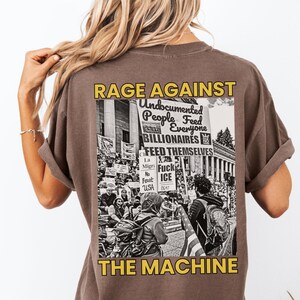 Puede incluir: Camiseta marr&oacute;n con un gr&aacute;fico en la espalda que dice "RAGE AGAINST THE MACHINE". El gr&aacute;fico incluye texto sobre personas indocumentadas, multimillonarios e ICE, junto con una imagen en blanco y negro de una protesta.
