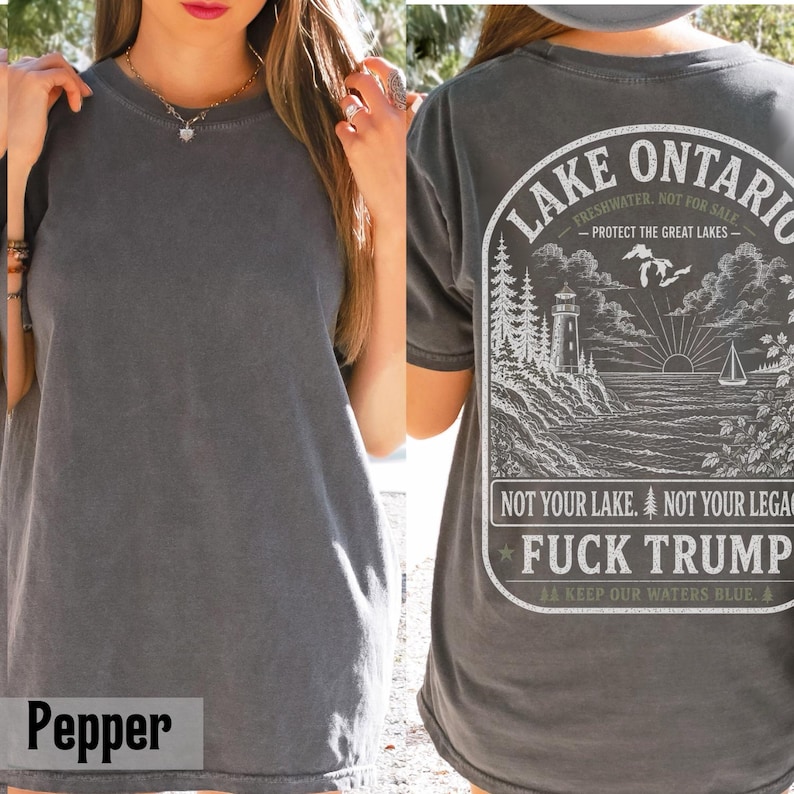 May include: A charcoal gray t-shirt featuring a white graphic on the back. The design depicts a lighthouse, a sailboat, and pine trees by a lake, with the text "LAKE ONTARIO," "PROTECT THE GREAT LAKES," "NOT YOUR LAKE. NOT YOUR LEGACY," and "FUCK TRUMP."