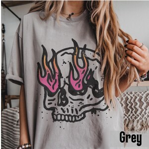May include: A grey t-shirt featuring a black skull graphic with pink and orange flames. The word "Grey" is printed in the bottom right corner of the image. The shirt has rolled-up sleeves.