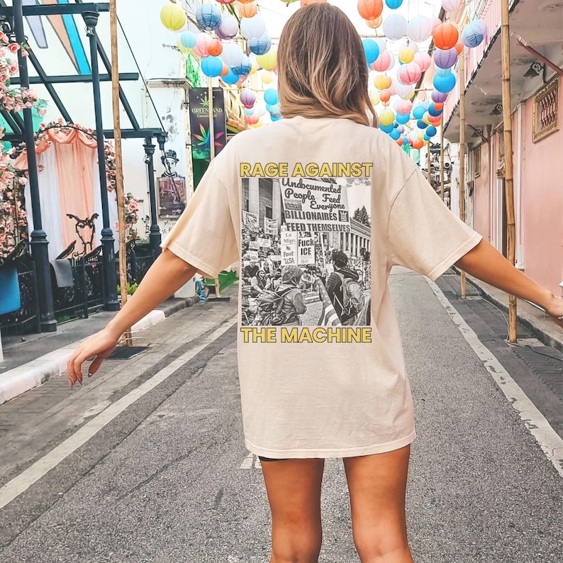 May include: Beige t-shirt with a graphic on the back that reads "RAGE AGAINST THE MACHINE." The graphic is a black and white image of a protest with text. The shirt is a relaxed fit.